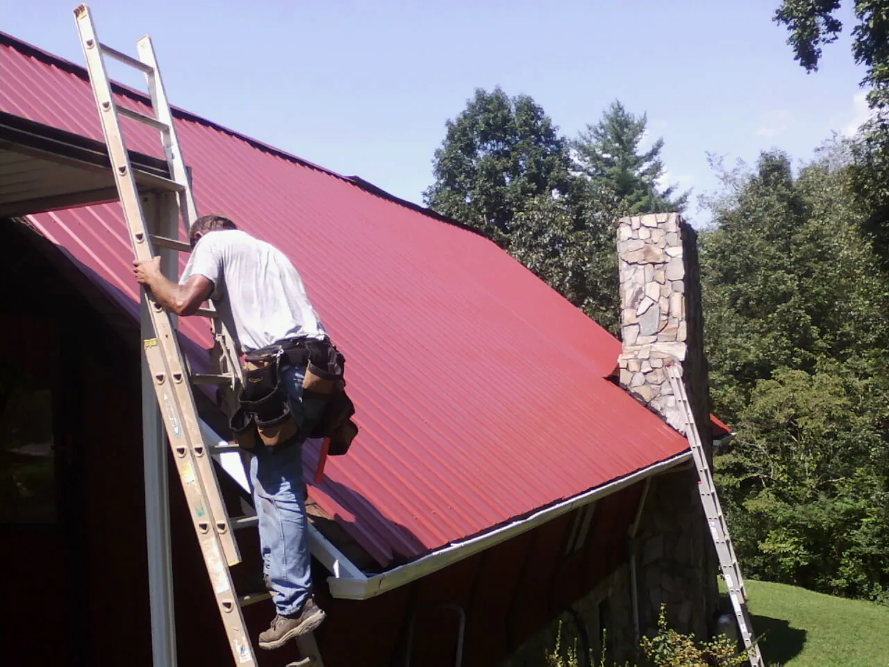 Roofer on a ladder beside a red metal roof and stone chimney — Soffit & Fascia Repair by Roofing Brothers in Arab
